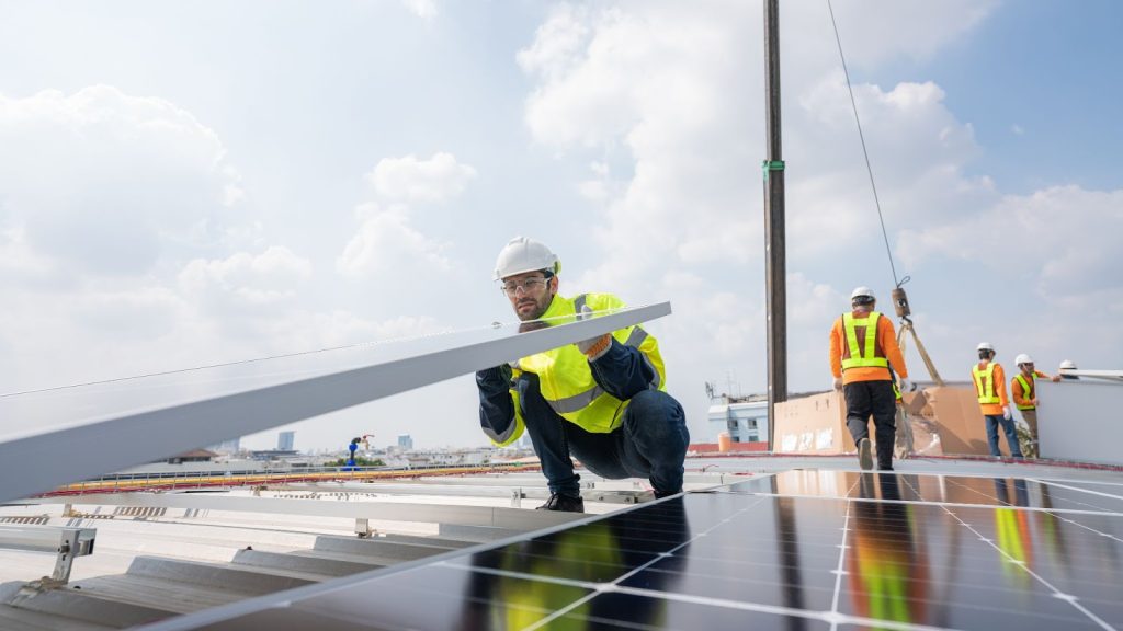 A man in a hard hat and safety vest installs solar panels on a new roof, preparing the home for energy efficiency.