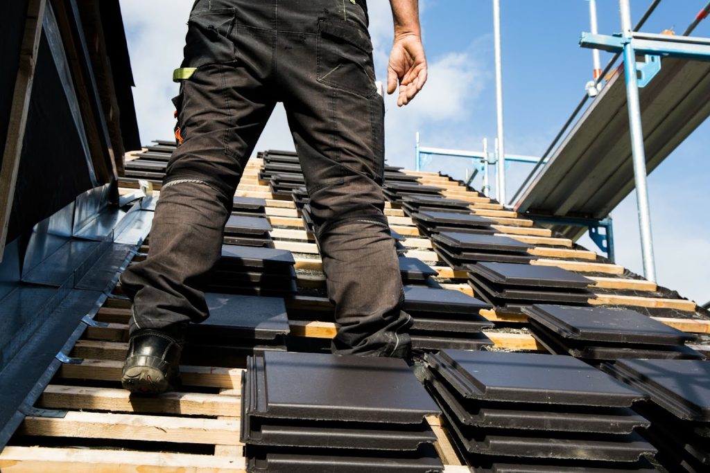 A man stands on a new roof, arranging a stack of black tiles as part of home preparation for roofing work.